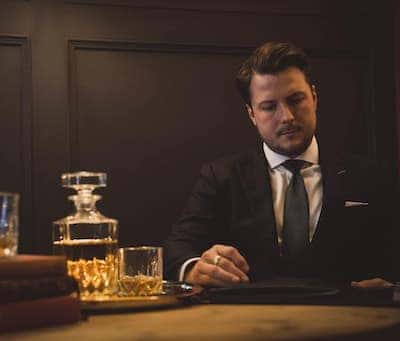 Man looking through fabric folder wearing black suit and navy tie next to whiskey bottle